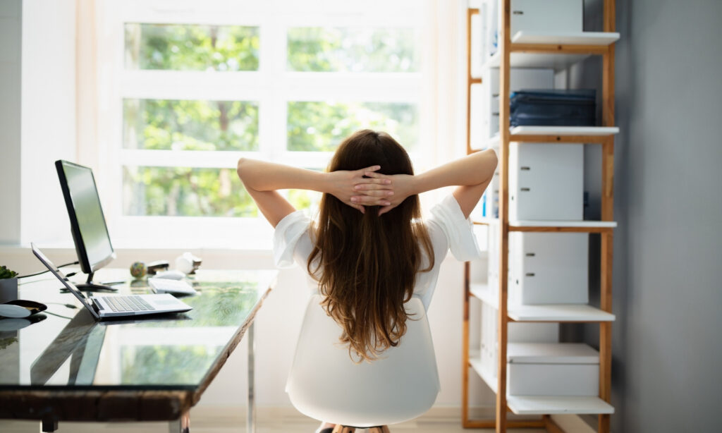 1. Mulher sentada em uma mesa de home office, com as mãos atrás da cabeça, demonstrando relaxamento e concentração.  2. Mulher em um home office, sentada à mesa com as mãos apoiadas atrás da cabeça, refletindo tranquilidade e foco.  3. Em um ambiente de home office, mulher sentada à mesa com as mãos atrás da cabeça, expressando calma e produtividade. O que é Home Office: Entenda Essa Modalidade de Trabalho