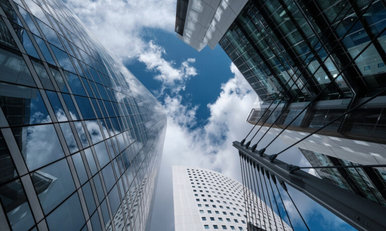 A imagem mostra um conjunto de arranha-céus modernos com fachadas de vidro refletindo o céu azul com nuvens. Essa perspectiva arquitetônica pode simbolizar o setor de fundos imobiliários, remetendo à valorização de grandes edifícios comerciais e a urbanização, elementos típicos de ativos imobiliários. Fundos imobiliários frequentemente investem em propriedades assim, que geram rendimentos através de aluguéis ou valorização. Fundos Imobiliários: Quais FIIs selecionar em outubro?