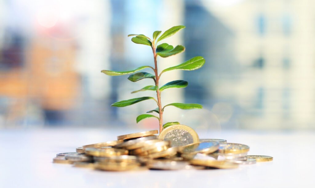 Planta jovem crescendo em uma pilha de moedas com fundo desfocado de paisagem urbana, simbolizando o investimento no setor rural. Micro-investimentos em Agricultura Urbana: Como Ganhar Dinheiro Cultivando na Cidade