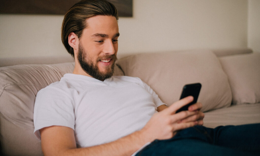 Pessoa de camisa branca sentada no sofá olhando para um smartphone.  1. Homem de barba, vestido com camisa branca, sentado no sofá, utilizando o celular enquanto busca dicas de renda extra.2. Homem barbudo em camisa branca, relaxando no sofá e consultando o celular para aprender sobre renda extra em horas livres.3. Homem com barba e camisa branca, sentado confortavelmente no sofá, usando o telefone para descobrir formas de renda extra.4. Homem de barba, usando uma camisa branca, sentado no sofá e acessando o celular em busca de dicas para ganhar renda extra.5. Homem barbudo em uma camisa branca, sentado no sofá, utilizando o celular para explorar opções de renda extra em seu tempo livre.  Como Ganhar Renda Extra em Horas Livres: Dicas Práticas Para Quem Tem Pouco Tempo