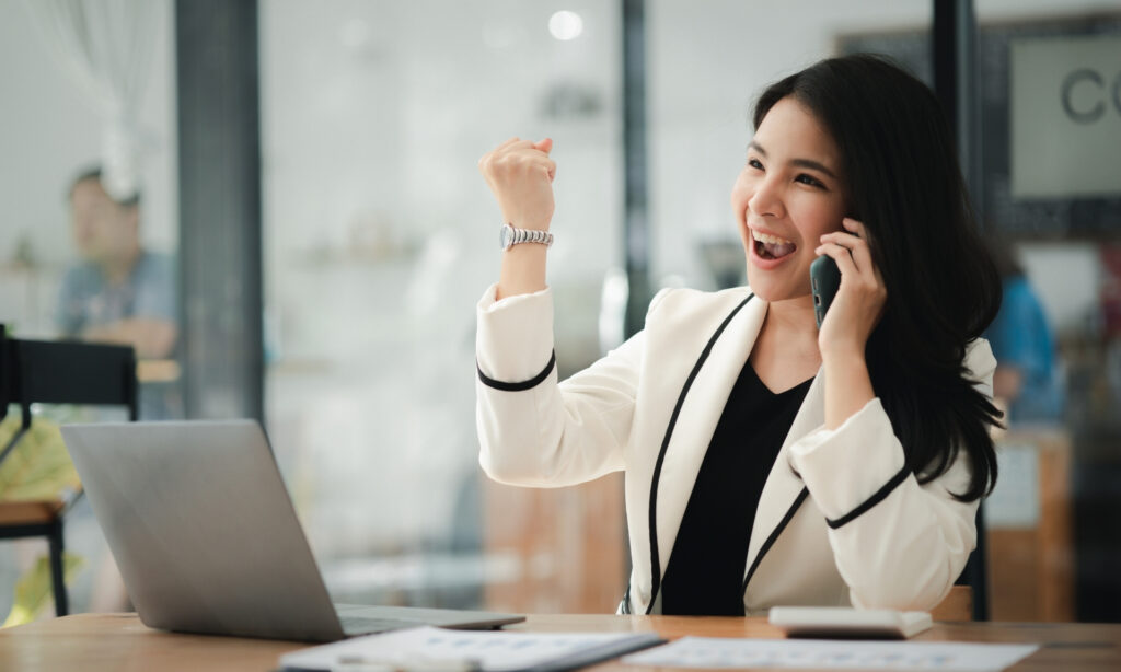 Uma pessoa de blazer branco sentada em uma mesa com o punho levantado, com um laptop e documentos na frente dela.  1. Mulher executiva em terno fala ao telefone, levantando o punho em sinal de determinação e confiança.  2. Executiva de conta em terno discute ao telefone, erguendo o punho em um gesto de assertividade.  3. Mulher em traje de negócios conversa ao telefone, levantando o punho em um gesto de empoderamento.  4. Executiva em terno fala ao telefone, levantando o punho como símbolo de força e liderança.  5. Mulher em traje profissional discute ao telefone, erguendo o punho em um gesto de motivação e firmeza.  Executivo de conta. 12 Profissões com Flexibilidade no Trabalho para um Equilíbrio Ideal