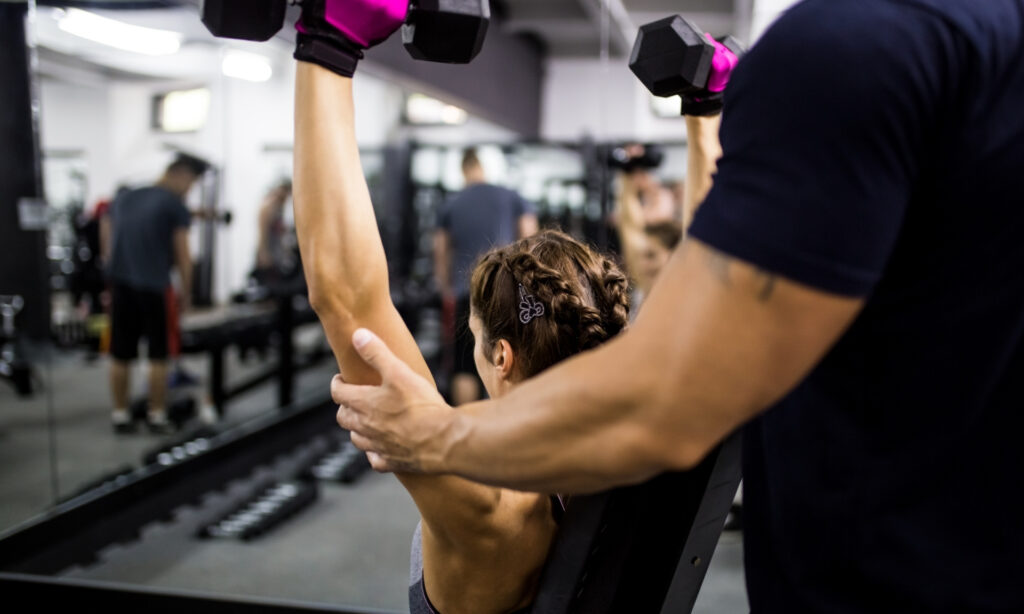 Mulher levantando halteres com a ajuda de um observador em uma academia.  1. Homem e mulher em uma academia, levantando pesos sob a orientação de um personal trainer.  2. Casal em uma sala de musculação, utilizando pesos com a supervisão de um personal trainer.  3. Homem e mulher treinando com pesos em uma academia, acompanhados por um personal trainer.  12 Profissões com Flexibilidade no Trabalho para um Equilíbrio Ideal