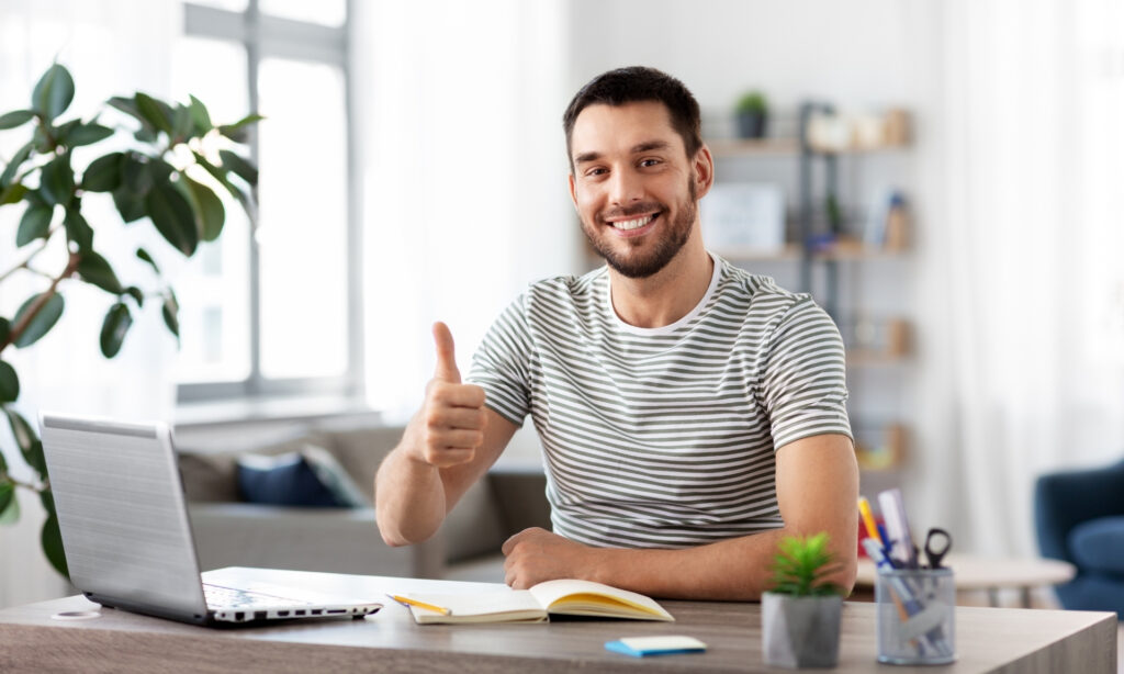 Como Ganhar Renda em Dólar e Proteger Seu Dinheiro da Alta da Moeda Homem sorridente mostrando sinal positivo enquanto trabalha remotamente em um ambiente bem iluminado, representando oportunidades de trabalho para empresas estrangeiras. Como Ganhar Renda em Dólar e Proteger Seu Dinheiro da Alta da Moeda